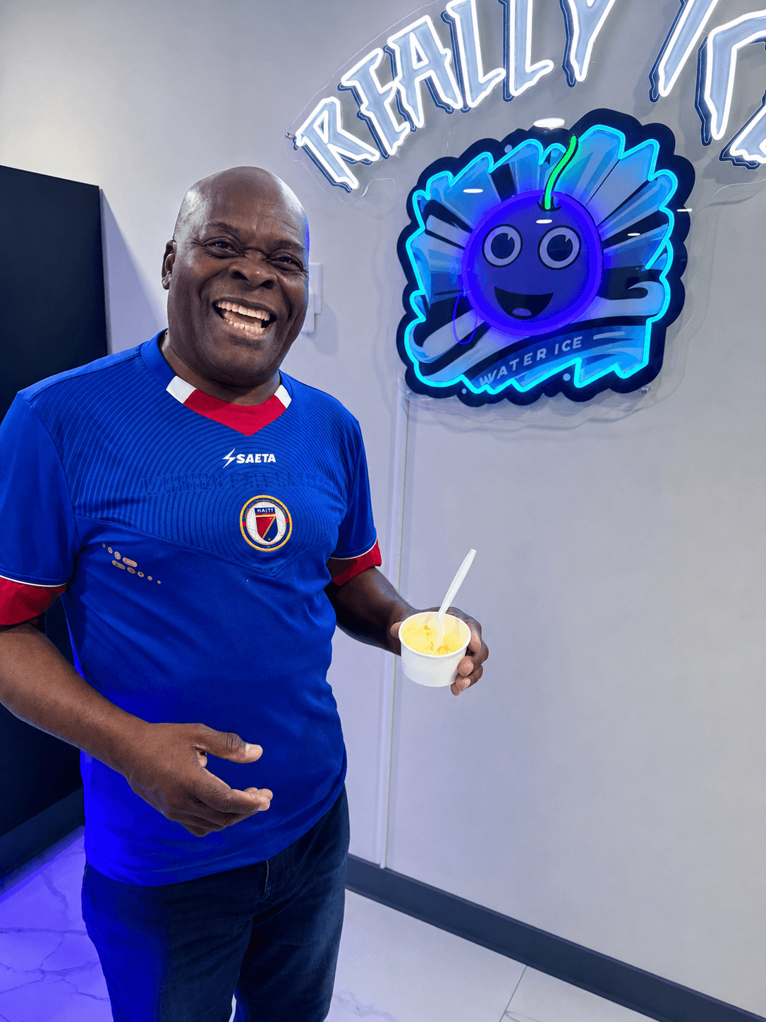 A smiling customer in a soccer jersey holding water ice under the Really Ice neon sign